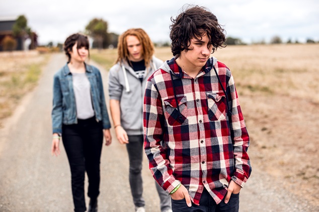 A young male teenager in a flannel shirt walking along a country road ahead of his two friends, who are speaking with each other.