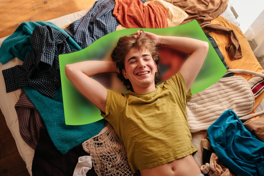A young person laying on their bed on a pile of clothes. The young person is smiling and laying on their back with their hands behind their head.
